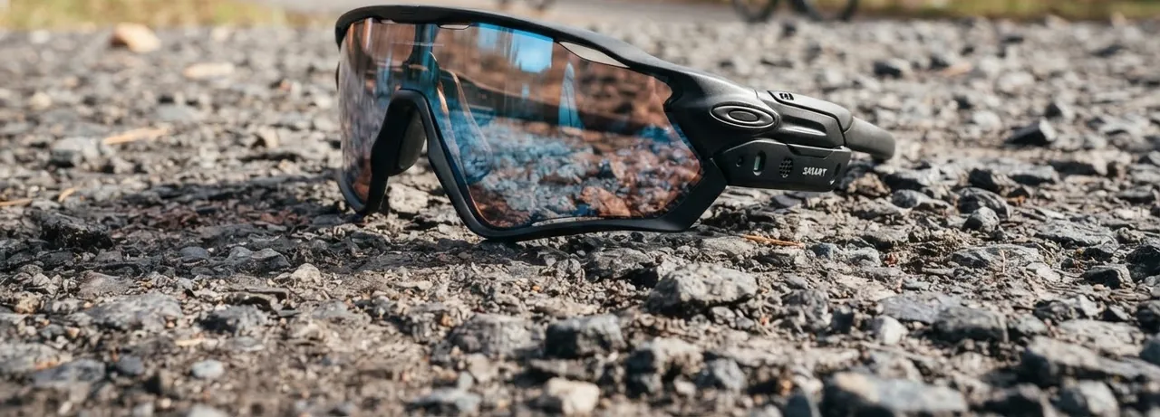 Gafas deportivas envolventes de lente espejada apoyadas sobre grava, con ciclistas desenfocados al fondo. (imagen generada con IA)